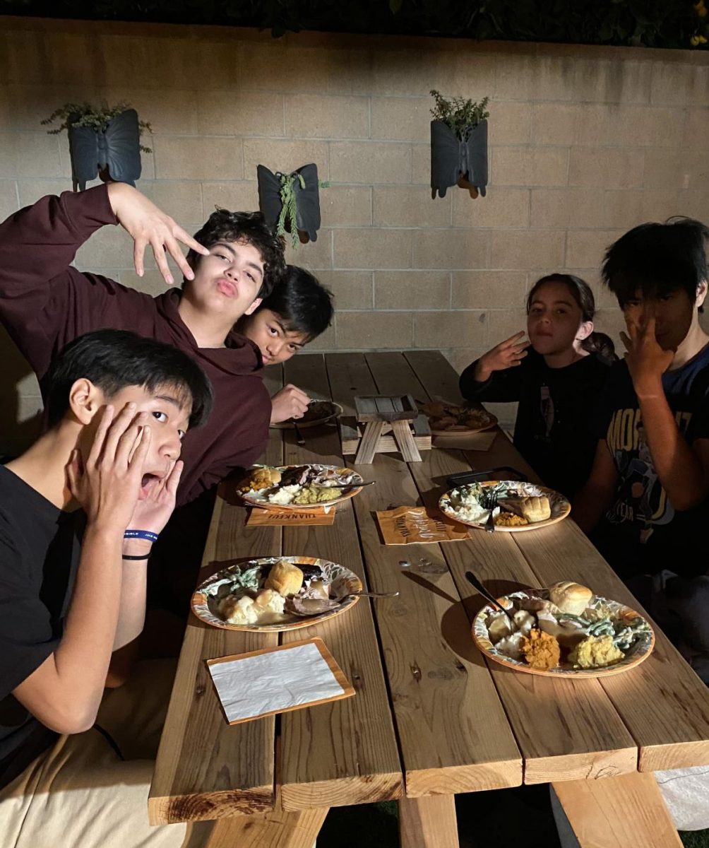 A group of friends sitting around a wooden picnic table outdoors at night enjoying a nice Thanksgiving meal.